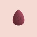 A Aspilon Cosmetics teardrop-shaped makeup sponge in a burgundy color centered on a solid pale pink background.